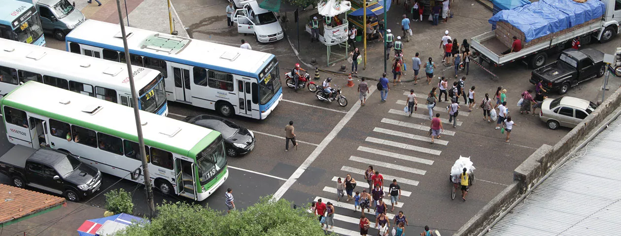 Foto mostra o cruzamento de uma avenida movimentada de cima, com a faixa de pedestre e ônibus e carros esperando no semáforo 