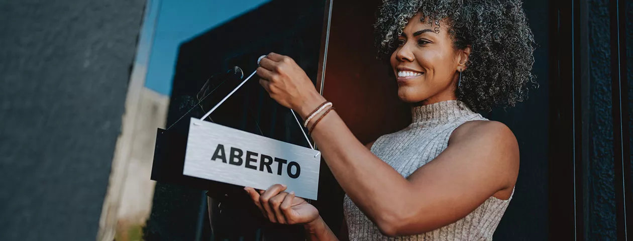 Foto mostra uma empreendedora colocando a placa "aberto" na frente do seu estabelecimento 