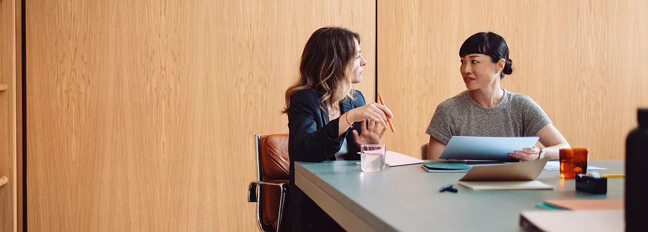 duas mulheres conversando em uma sala de reunião enquanto anotam algo. 