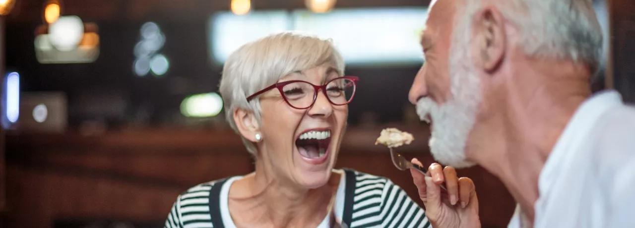 A imagem mostra um casal com cabelos grisalhos se alimentando em um restaurante 