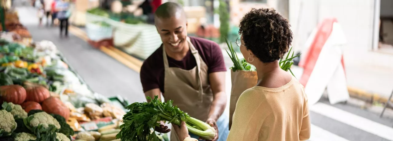 A imagem mostra uma mulher comprando legumes na feira, com um vendedor atendendo ela 