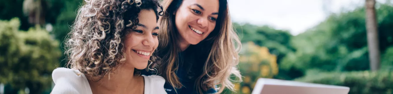 duas mulheres sorrindo enquanto olham algo em computador ao ar livre. 