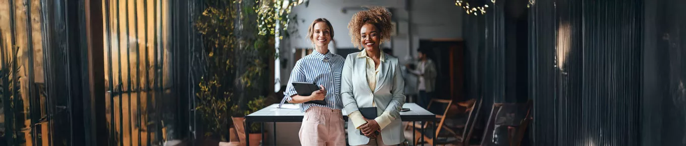 duas mulheres em ambiente corporativo segurando cadernos sorrindo enquanto olham para a câmera. 