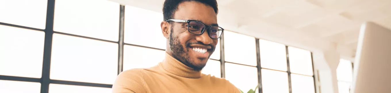 Homem negro, de camiseta amarela olhando para o notebook e sorrindo 