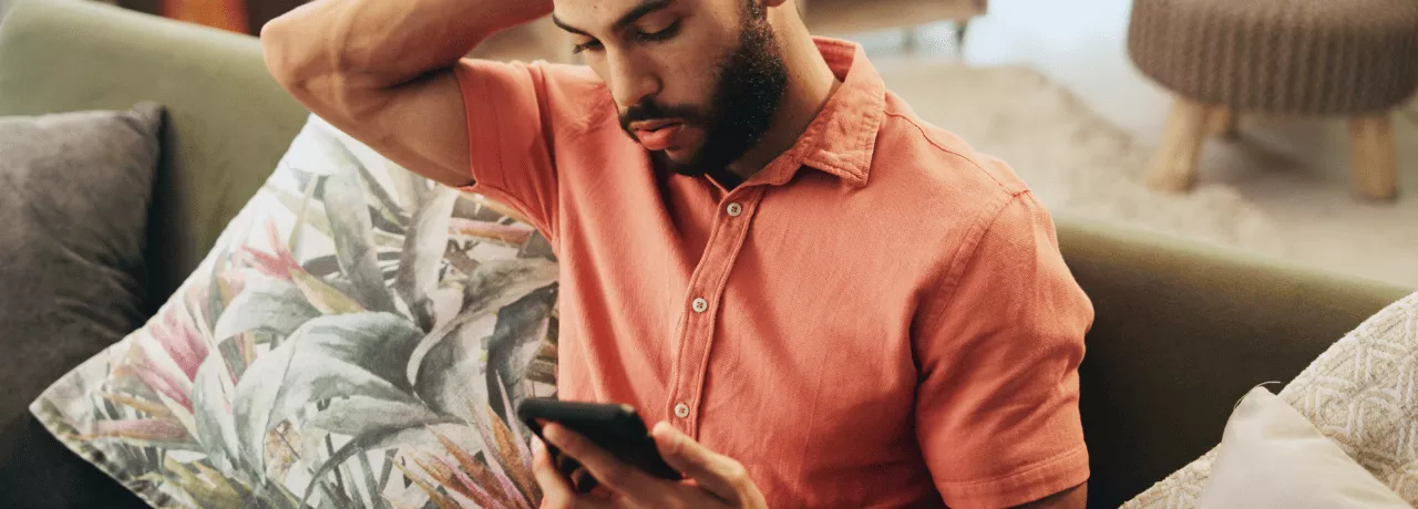 Pessoa sentada em um sofá, usando uma camisa de cor salmão, olhando para um smartphone que segura com as duas mãos. Ao lado há almofadas decorativas, incluindo uma com estampa de folhas. O ambiente interno é aconchegante, com iluminação suave e móveis desfocados ao fundo. 
