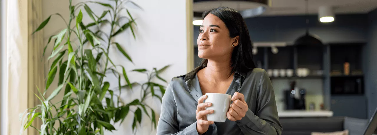 A imagem mostra uma mulher, em casa, segurando uma caneca e sorrindo olhando para a janela