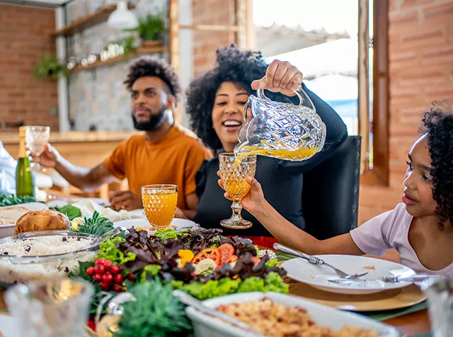 Foto mostra almoço de família. A mãe está servindo suco de laranja para sua filha.