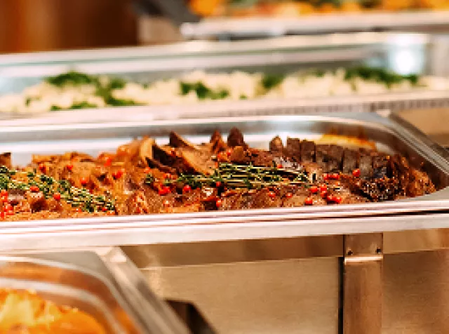 Buffet com pratos variados em réchauds de inox, incluindo carnes assadas e acompanhamentos