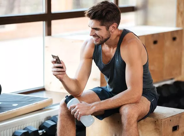 Pessoa sentada em área de treino segurando um celular e uma garrafa de água.