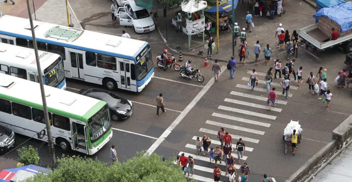 Foto mostra o cruzamento de uma avenida movimentada de cima, com a faixa de pedestre e ônibus e carros esperando no semáforo