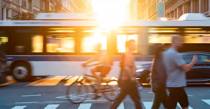 Foto mostra um cruzamento de avenida, com pedestres passando na faixa e um ônibus de fundo