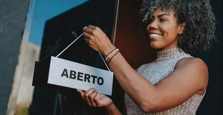Foto mostra uma empreendedora colocando a placa "aberto" na frente do seu estabelecimento