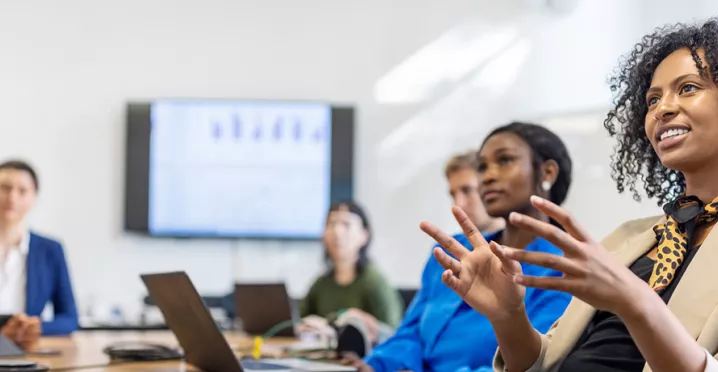 mulher explicando algo para uma sala de reunião com várias pessoas e apresentação ao fundo.