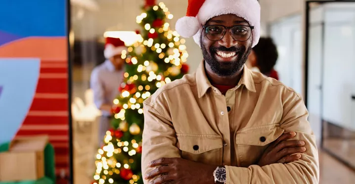 homem com chapéu de papai noel sorrindo na empresa com árvore de natal ao fundo.
