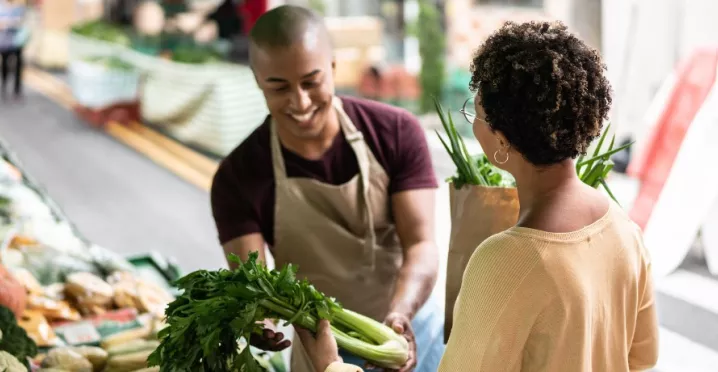A imagem mostra uma mulher comprando legumes na feira, com um vendedor atendendo ela