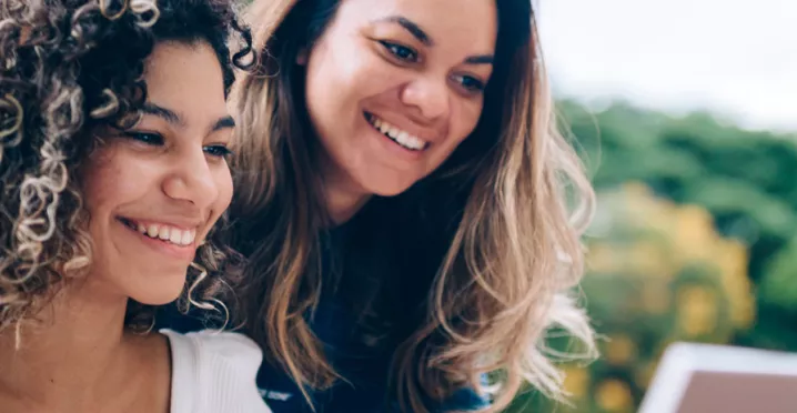 duas mulheres sorrindo enquanto olham algo em computador ao ar livre.