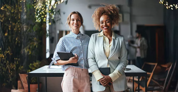 duas mulheres em ambiente corporativo segurando cadernos sorrindo enquanto olham para a câmera.