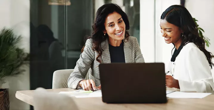 duas mulheres sorrindo enquanto conversam em frente a um notebook no escritório.