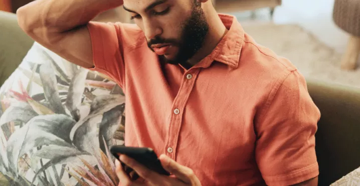 Pessoa sentada em um sofá, usando uma camisa de cor salmão, olhando para um smartphone que segura com as duas mãos. Ao lado há almofadas decorativas, incluindo uma com estampa de folhas. O ambiente interno é aconchegante, com iluminação suave e móveis desfocados ao fundo.