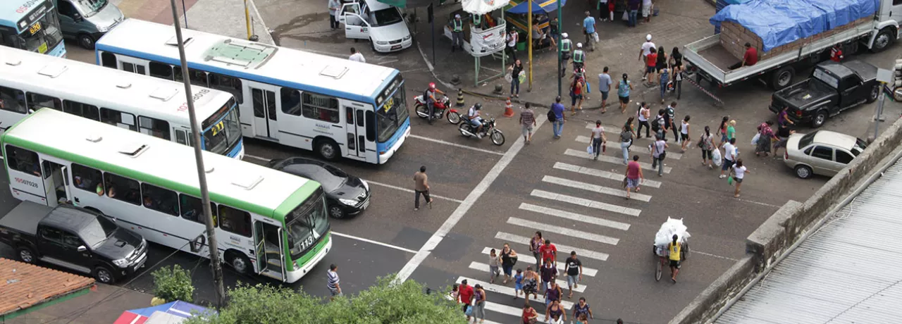 Foto mostra o cruzamento de uma avenida movimentada de cima, com a faixa de pedestre e ônibus e carros esperando no semáforo