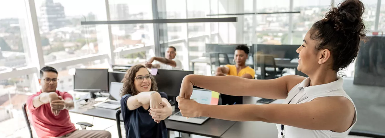 Quatro trabalhadores fazem ginástica laboral no escritório com uma professora