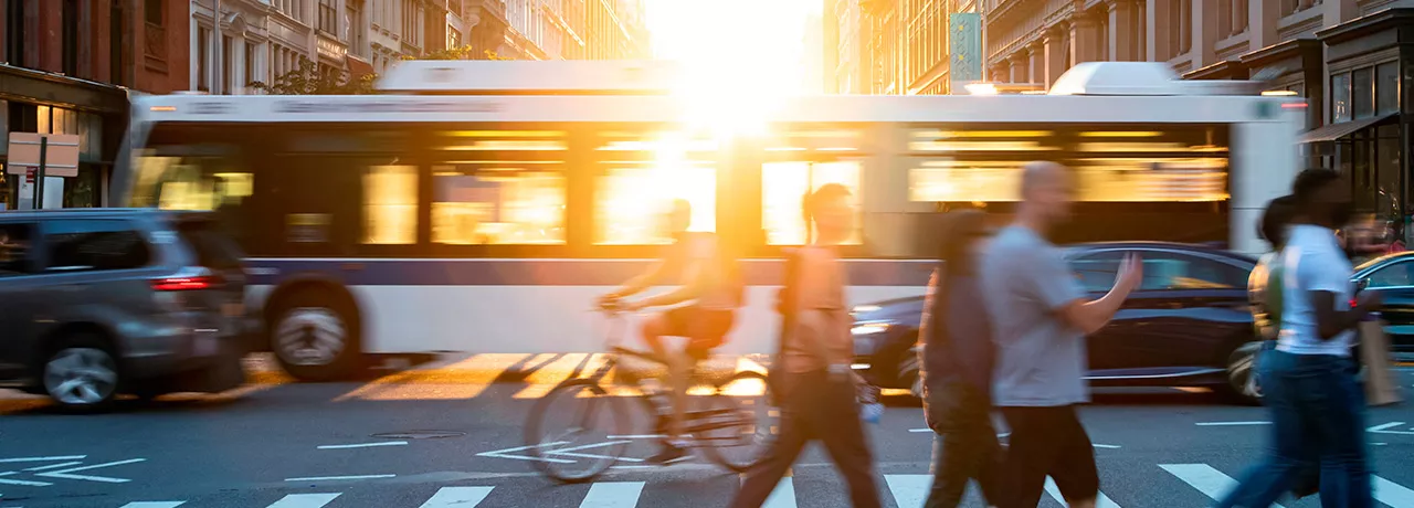 Foto mostra um cruzamento de avenida, com pedestres passando na faixa e um ônibus de fundo