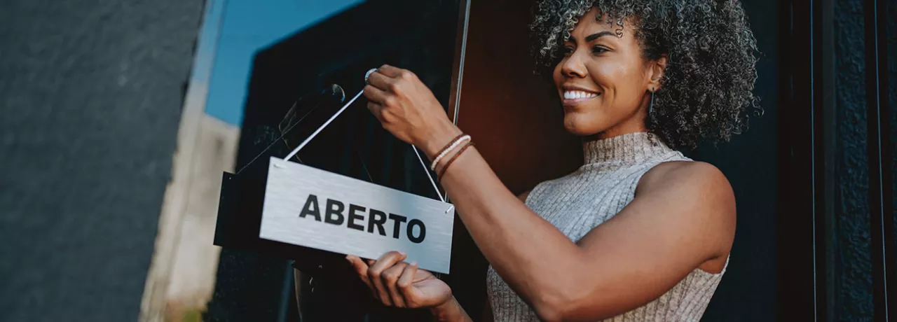 Foto mostra uma empreendedora colocando a placa "aberto" na frente do seu estabelecimento
