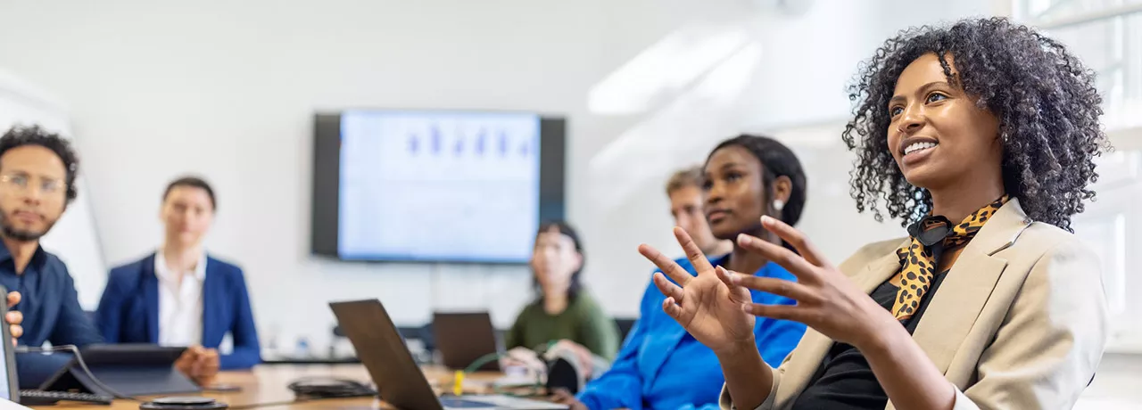 mulher explicando algo para uma sala de reunião com várias pessoas e apresentação ao fundo.