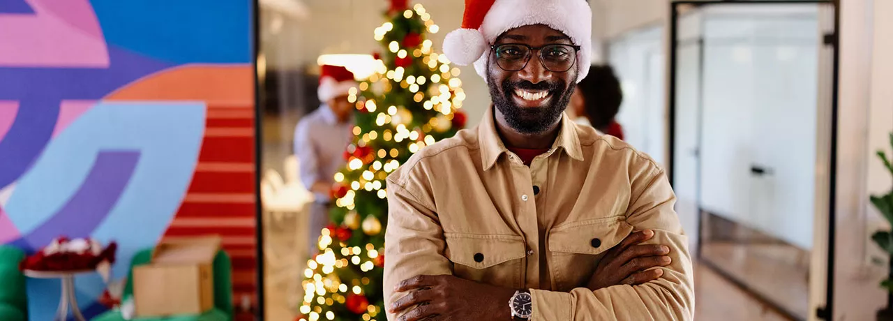 homem com chapéu de papai noel sorrindo na empresa com árvore de natal ao fundo.