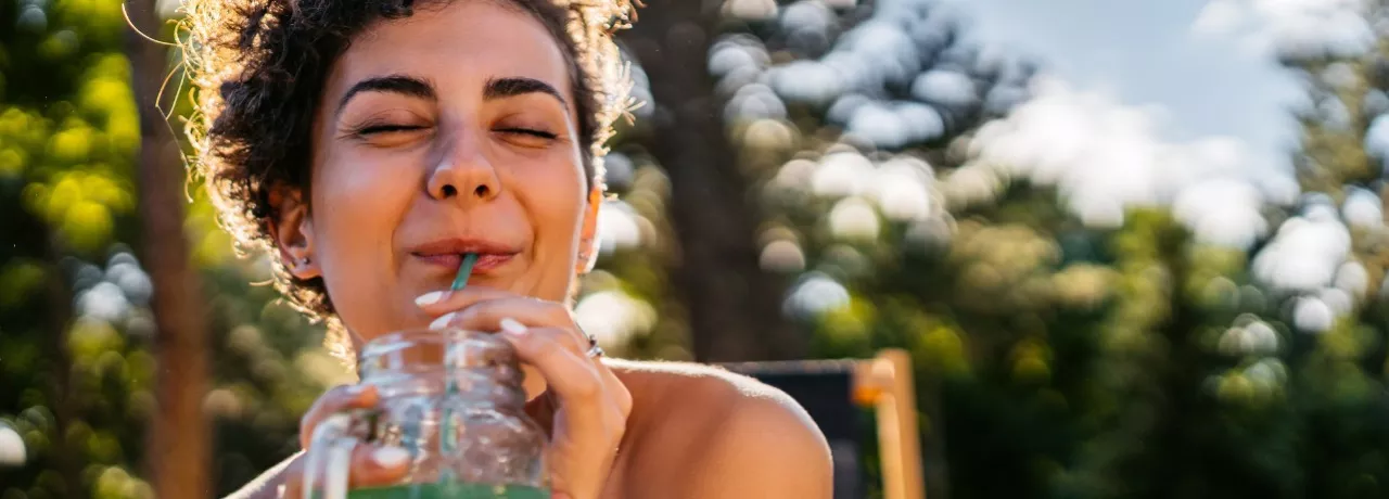 A imagem mostra uma mulher tomando suco enquanto toma um banho de sol.