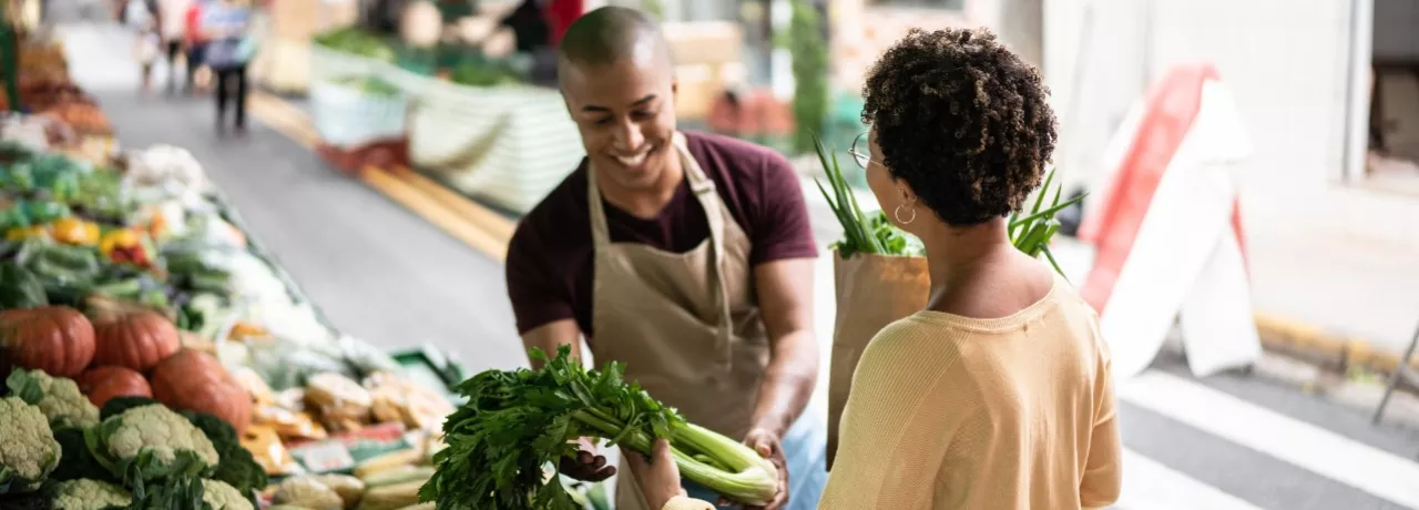 A imagem mostra uma mulher comprando legumes na feira, com um vendedor atendendo ela