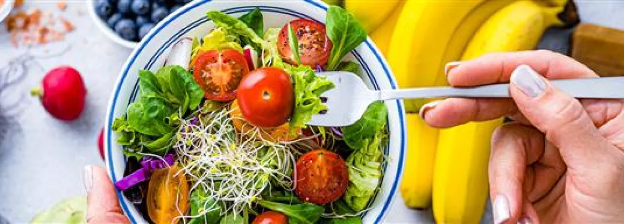 Imagem mostra uma salada com tomates e frutas ao fundo