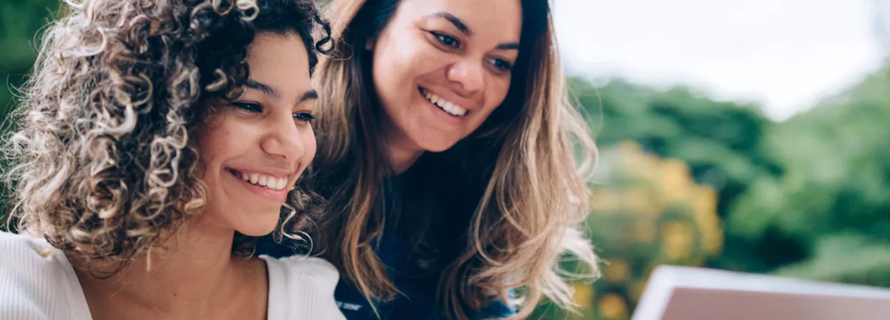 duas mulheres sorrindo enquanto olham algo em computador ao ar livre.