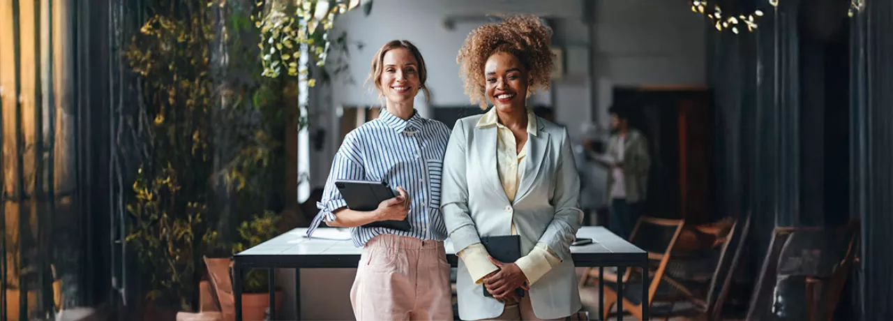 duas mulheres em ambiente corporativo segurando cadernos sorrindo enquanto olham para a câmera.