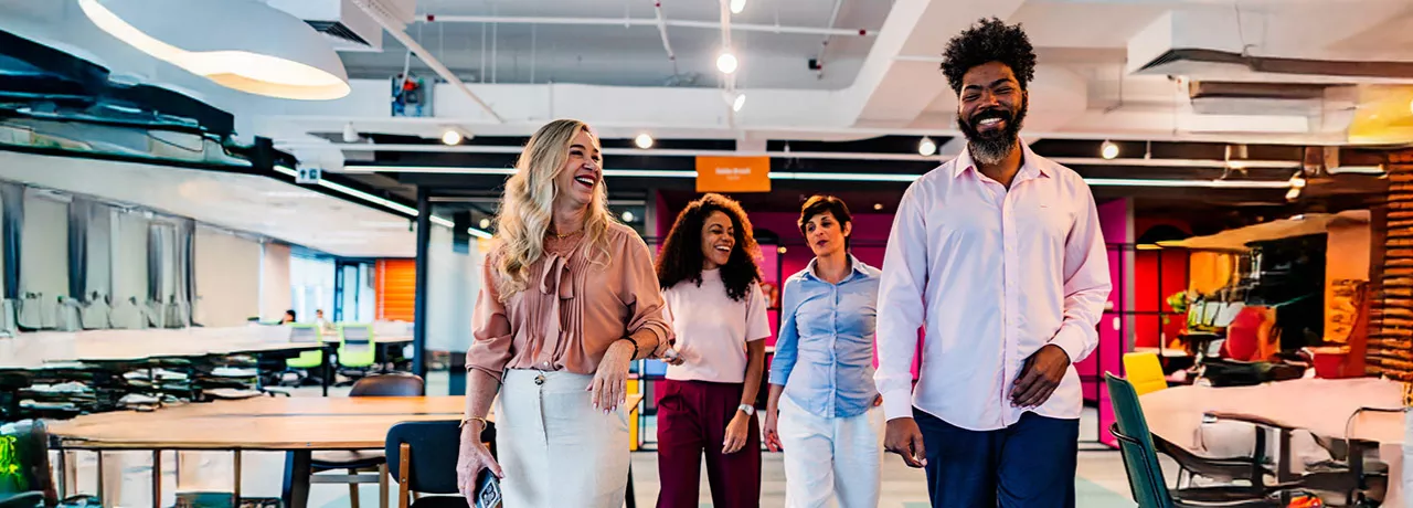 time de três mulheres e um homem caminhando, conversando e rindo em escritório.