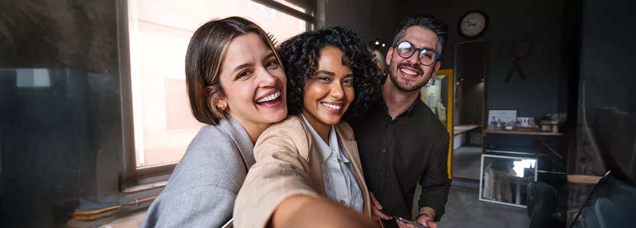 Foto mostra três colegas de trabalho tirando uma selfie no escritório