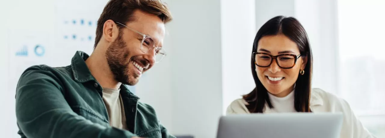homem e mulher, ambos de óculos, sorrindo em um ambiente corporativo