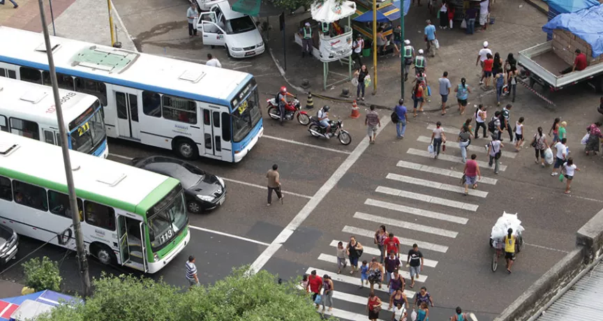 Foto mostra o cruzamento de uma avenida movimentada de cima, com a faixa de pedestre e ônibus e carros esperando no semáforo