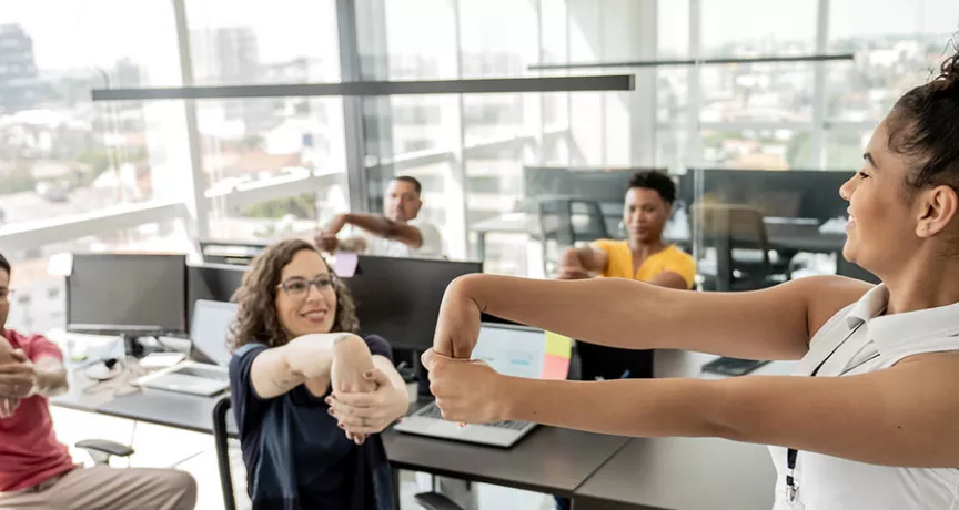 Quatro trabalhadores fazem ginástica laboral no escritório com uma professora