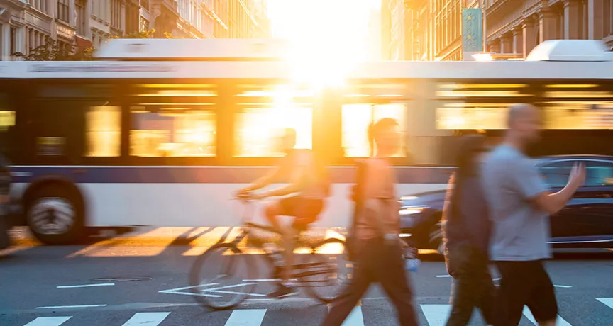 Foto mostra um cruzamento de avenida, com pedestres passando na faixa e um ônibus de fundo