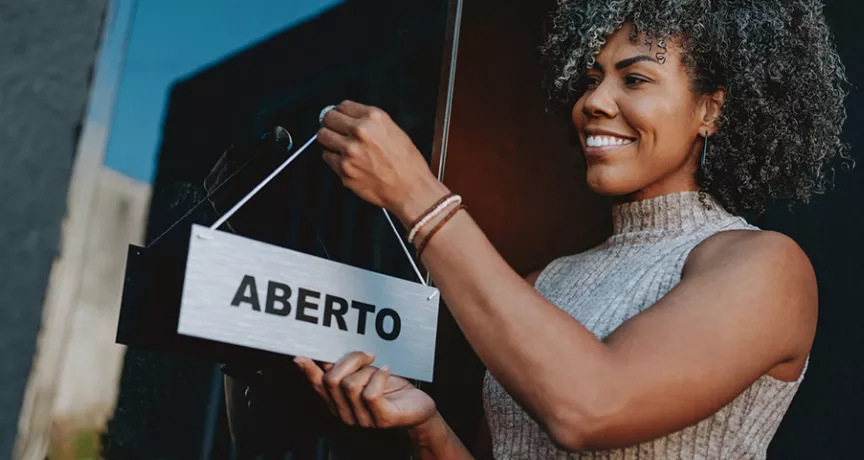 Foto mostra uma empreendedora colocando a placa "aberto" na frente do seu estabelecimento