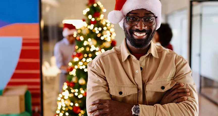 homem com chapéu de papai noel sorrindo na empresa com árvore de natal ao fundo.