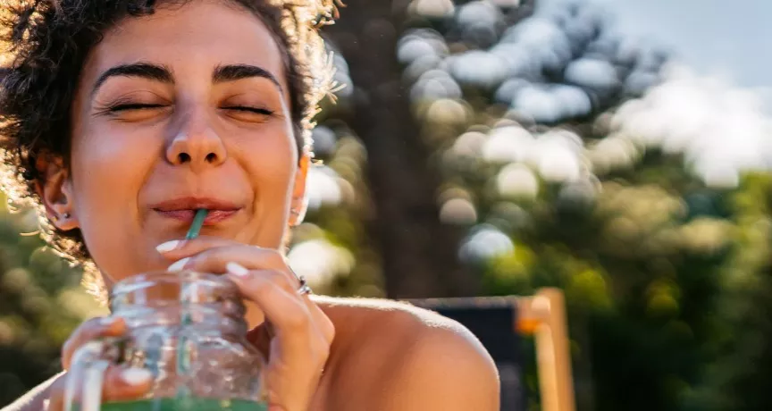 A imagem mostra uma mulher tomando suco enquanto toma um banho de sol.