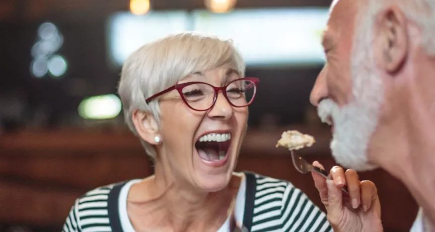 A imagem mostra um casal com cabelos grisalhos se alimentando em um restaurante
