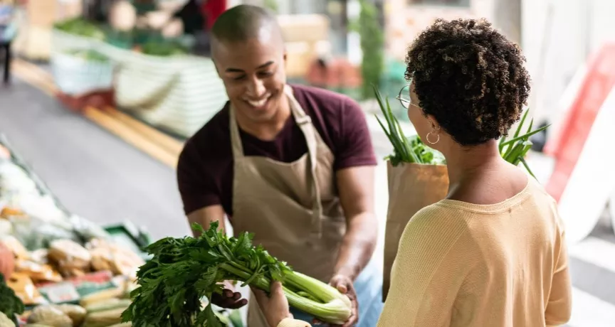 A imagem mostra uma mulher comprando legumes na feira, com um vendedor atendendo ela