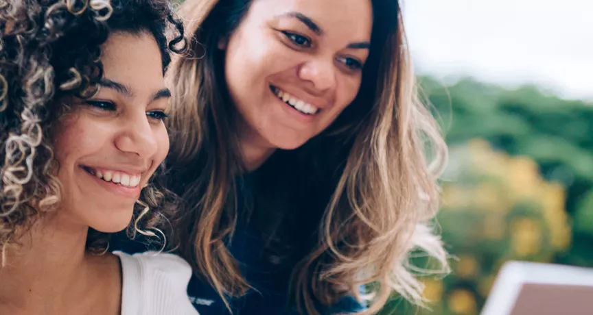 duas mulheres sorrindo enquanto olham algo em computador ao ar livre.