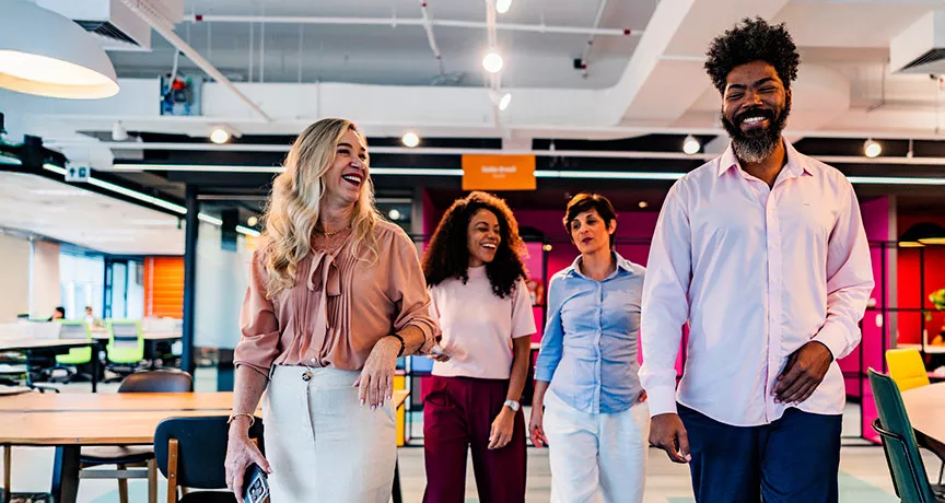 time de três mulheres e um homem caminhando, conversando e rindo em escritório.