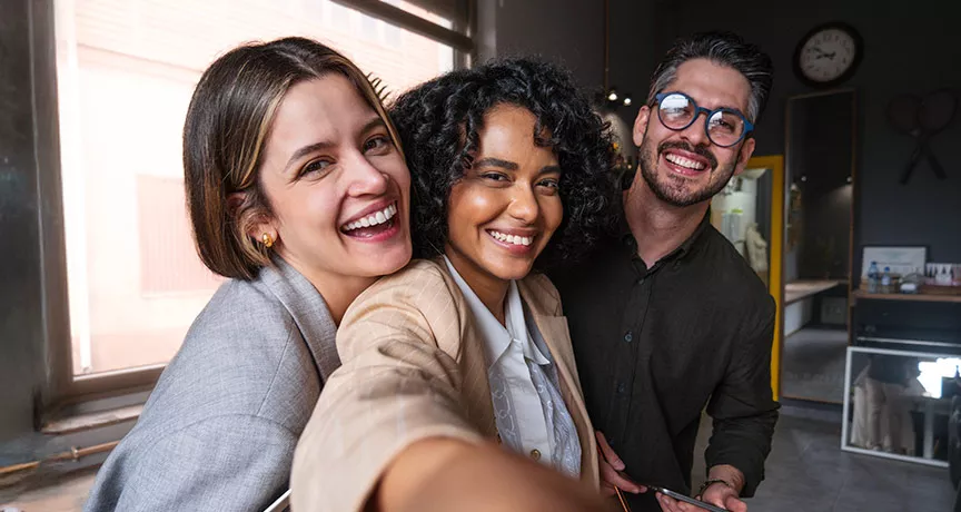 Foto mostra três colegas de trabalho tirando uma selfie no escritório