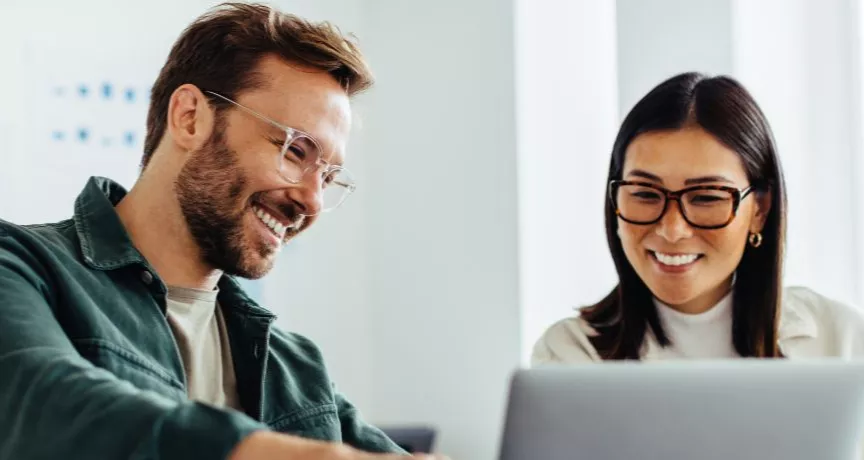 homem e mulher, ambos de óculos, sorrindo em um ambiente corporativo