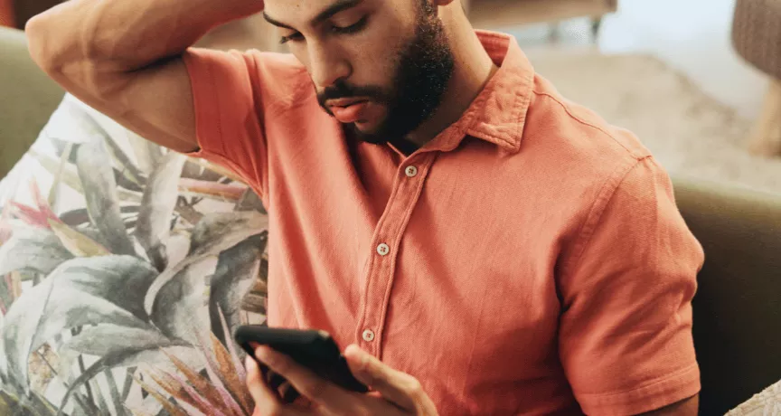 Pessoa sentada em um sofá, usando uma camisa de cor salmão, olhando para um smartphone que segura com as duas mãos. Ao lado há almofadas decorativas, incluindo uma com estampa de folhas. O ambiente interno é aconchegante, com iluminação suave e móveis desfocados ao fundo.
